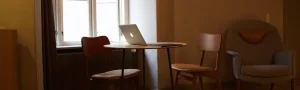 A MacBook sitting a table with two empty chairs. Behind is a big window, illuminating the dim room.