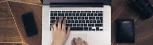 A person typing on their laptop, on a wooden table beside a cell phone, wallet, and camera.