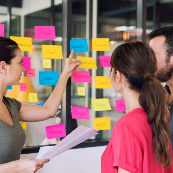 A range of sticky notes are attached to a glass panel. Three people standing nearby talk about what is printed on the notes.