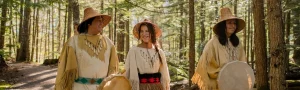 Three Indigenous people wearing traditional Indigenous clothing and walking through the forest on a sunny day.