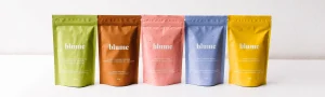 A product photo featuring bags of Blume latte blends in rainbow hues lined up on a white counter.