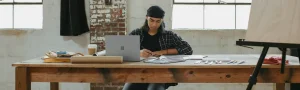 An artist working on a drawing with a laptop open on their desk.