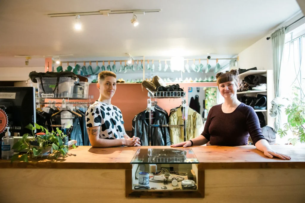 Awl Together Leather co-owners, Tess and Ariss, smiling and standing at a counter in their shop, with racks of jackets and shoes behind them.