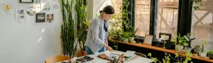 A person working on an art piece in a very bright, white studio space. Their space is filled with art, art supplies, and plants.