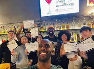 Selfie of a Fine Art Bartending instructor with their class. The wall behind them is filled with bottles.