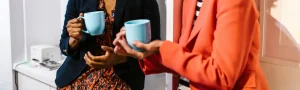 Two people in a bright office space laughing while holding coffee mugs.