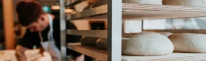 A baker works on bread while loafs of bread proof on racks.