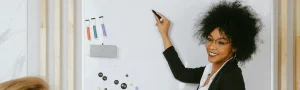 An instructor holds a pen up to a whiteboard while smiling at the room.