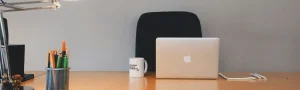 An open laptop sitting on a wooden desk beside a coffee mug, notebook, and pencils.