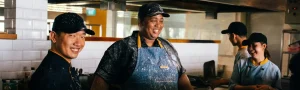 A team of bakers laugh and look happy while working in a kitchen.