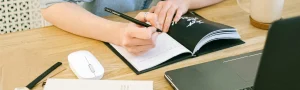 A person writes in a notebook on a desk in front of a laptop.