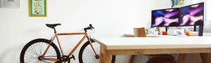 A bright home office with a large desk and computer monitors and a bicycle leaning against the wall.